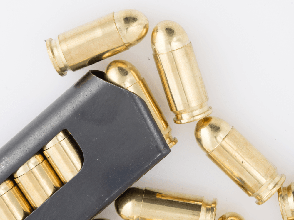 Cartuchos de acabamento dourado e formato arredondado dispostos ao redor de um carregador preto, fotografados sobre fundo branco.