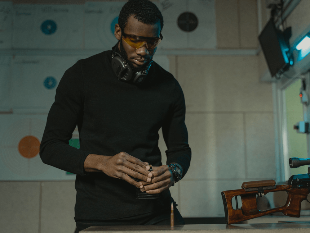 Homem usando óculos de proteção examina um cartucho ao lado de um rifle com coronha de madeira em ambiente de tiro esportivo. Tudo para explicar os tipos de munição.